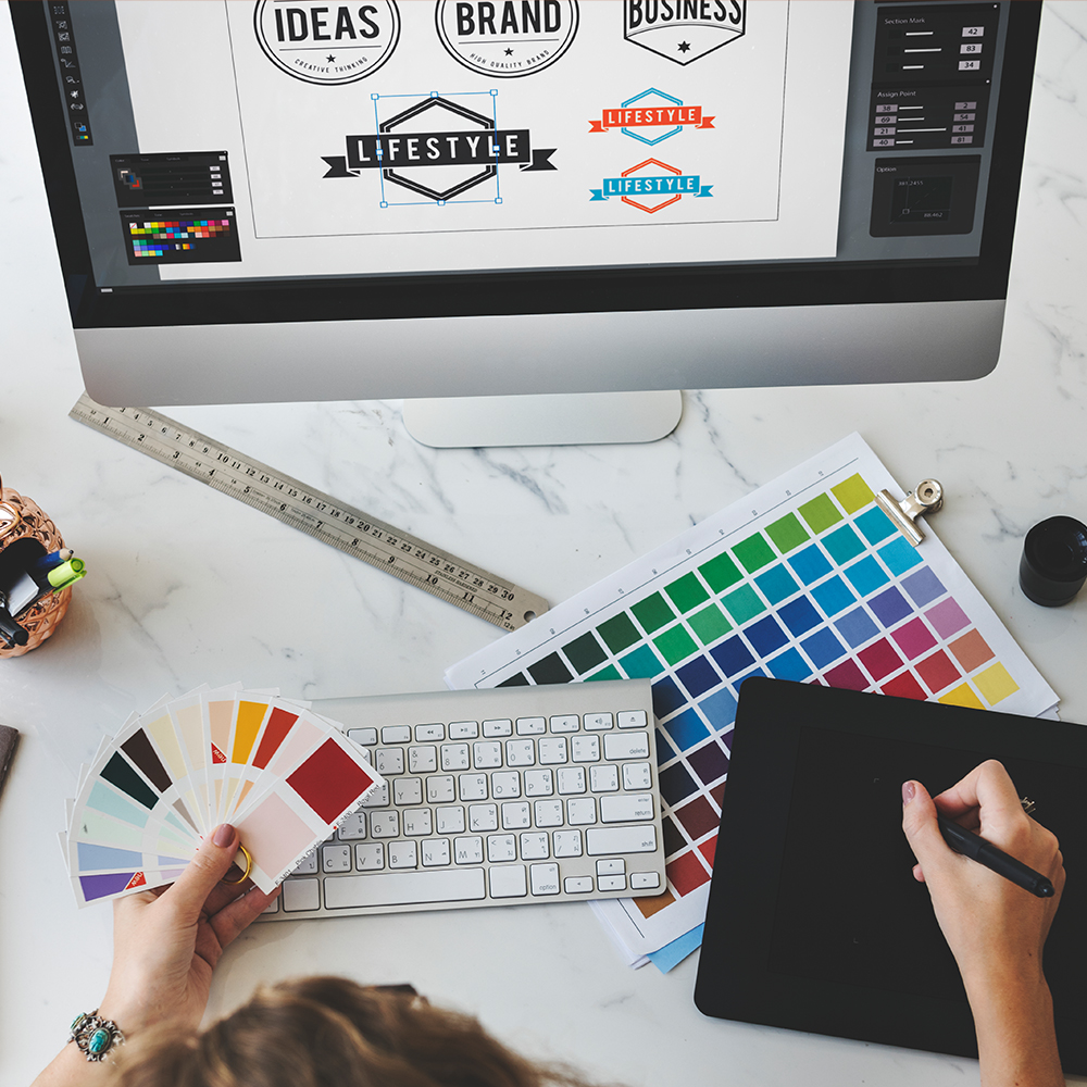 Top view of graphic designer working at their workstation holding color samples and using a graphics tablet with logo ideas on the screen of their computer.