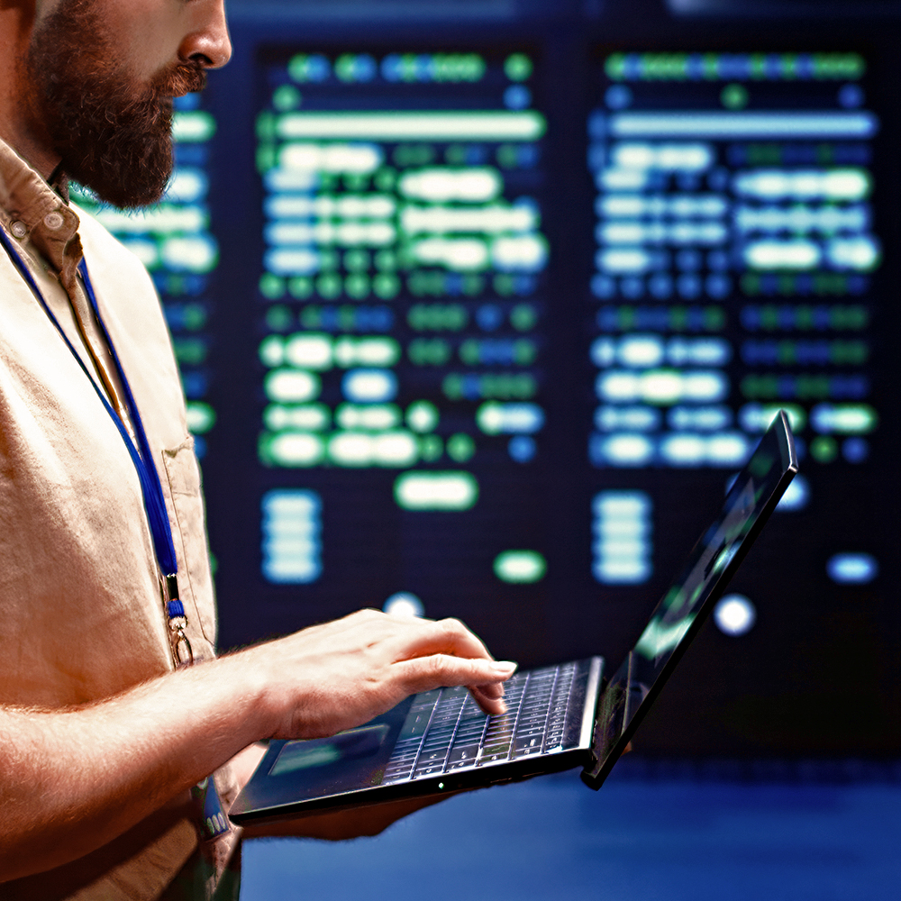 Technician wearing a lanyard holding a laptop while typing in front of a room full of computer servers